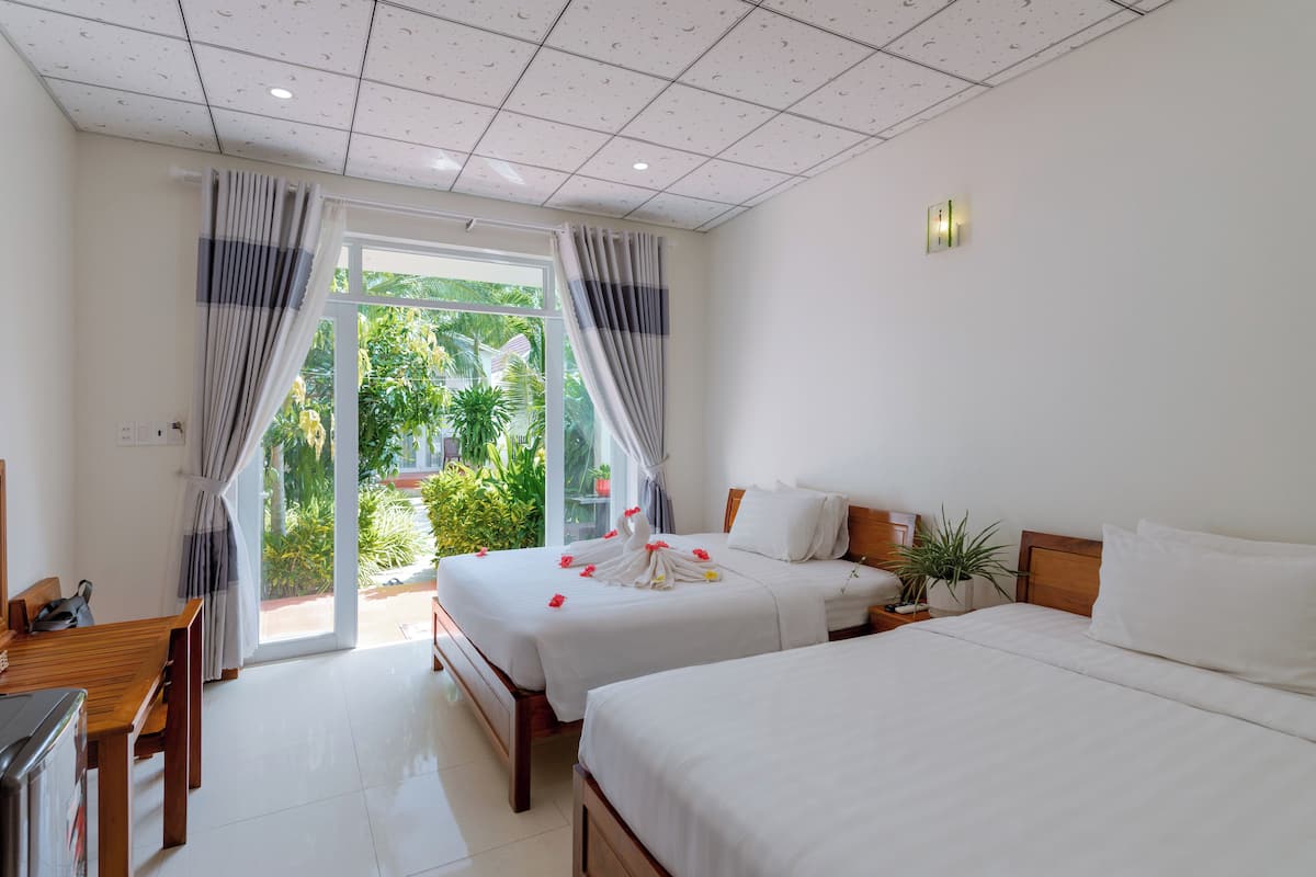 A hotel room with two beds, a wooden table, a TV, and a view of greenery through the window.