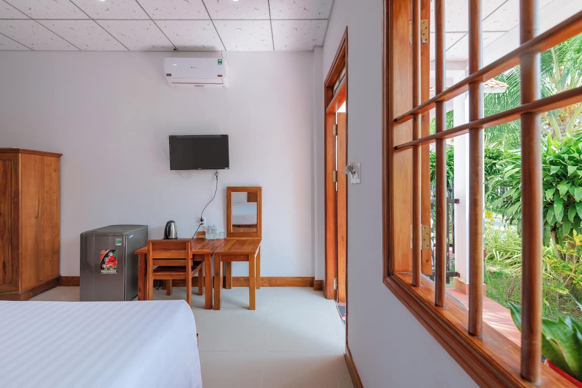 A hotel room with a bed, a desk with a chair, a television, and a window with a view of greenery.