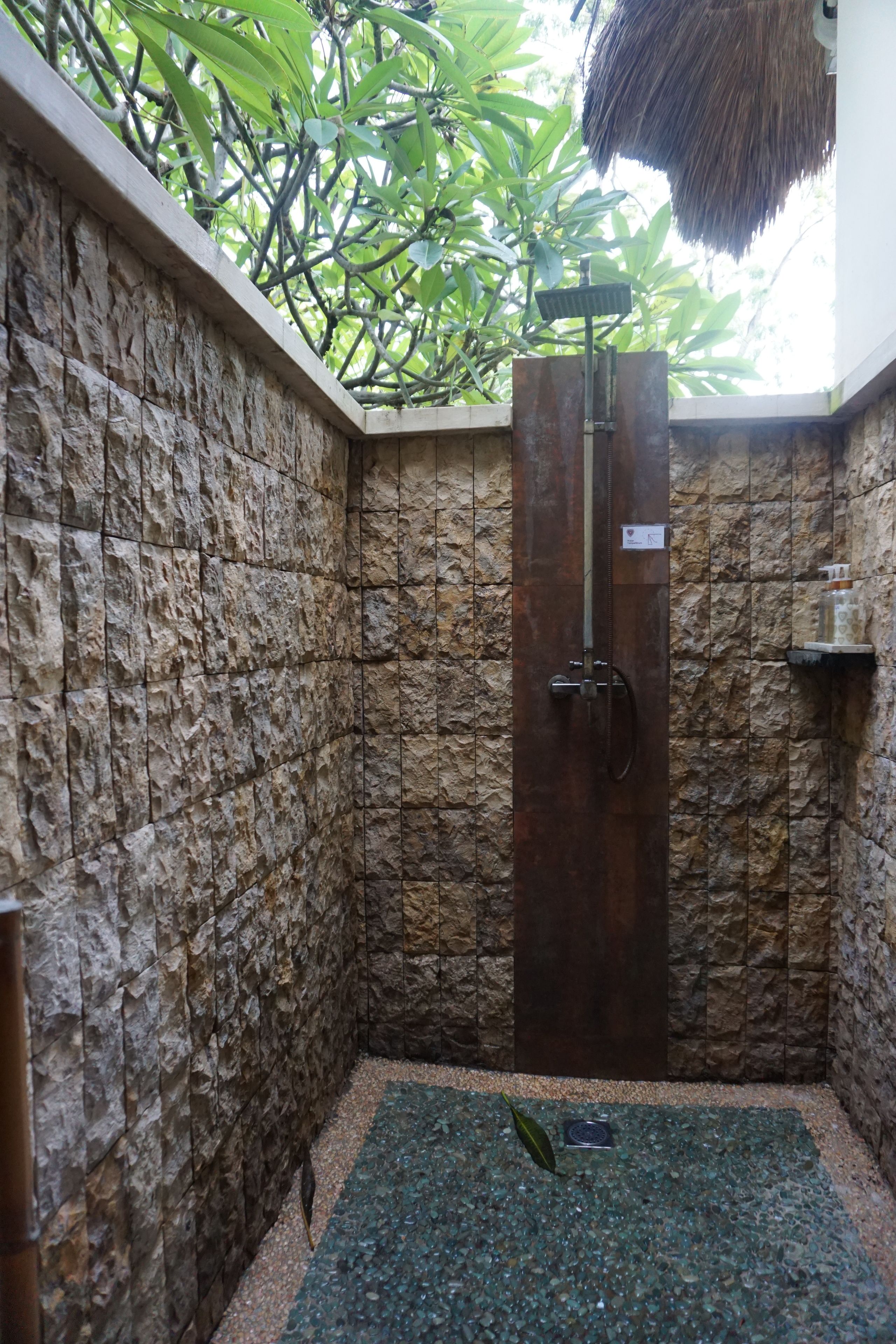 Une douche aux murs en pierre, dotée d’une porte en verre, d’une petite étagère et d’un toit en chaume.