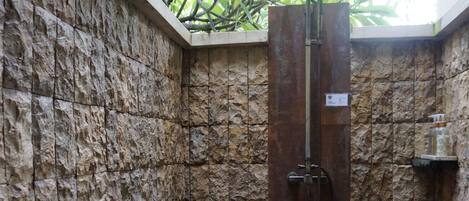 Una ducha con paredes de piedra, puerta de vidrio, un pequeño estante y techo de paja.