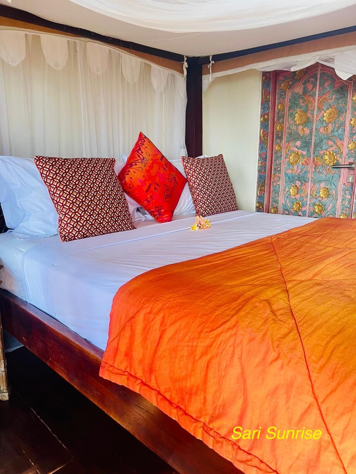 Una cama con ropa de cama blanca y naranja, almohadas estampadas en rojo y negro, y una cortina con motivos florales.
