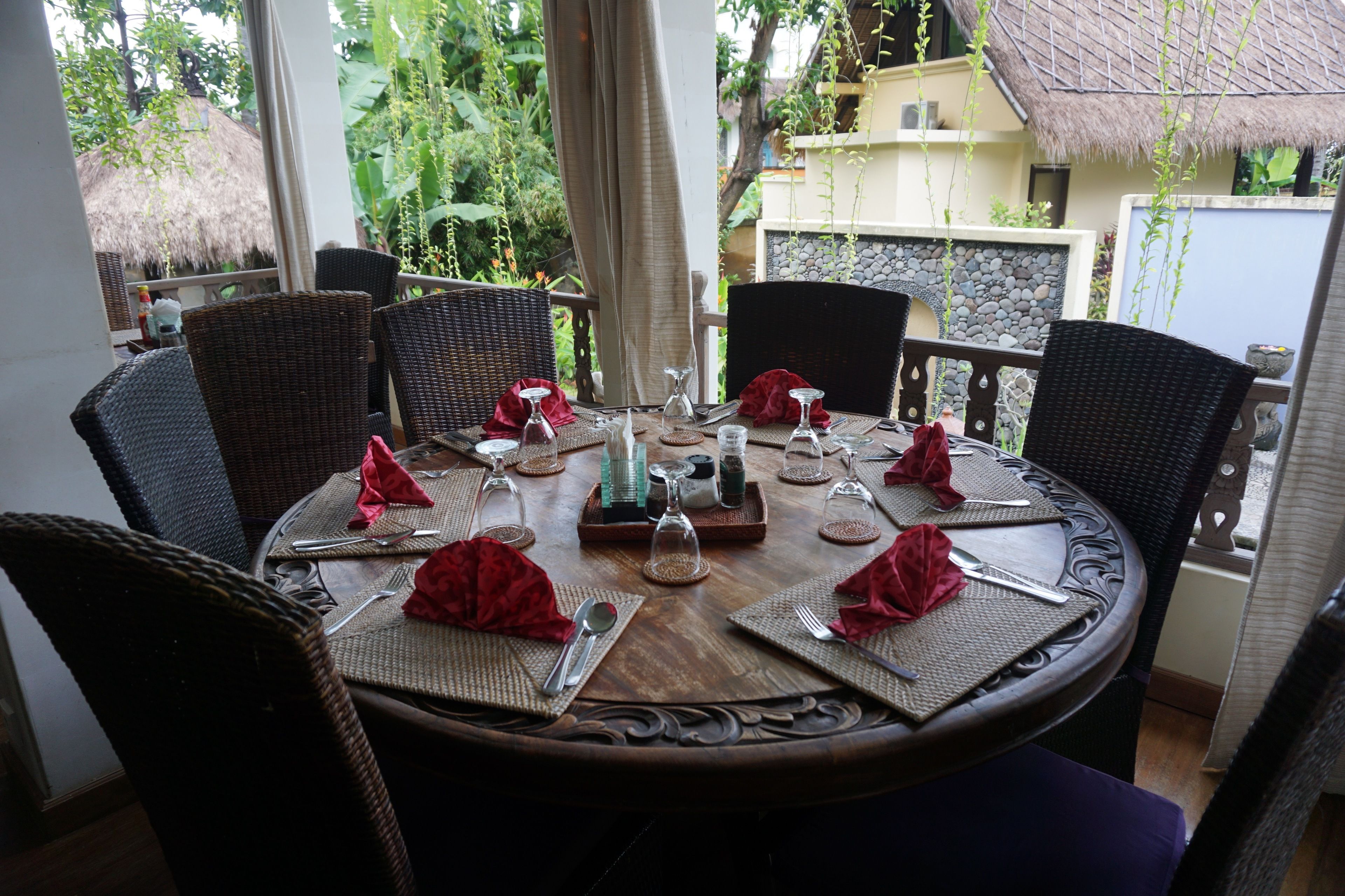 A round dining table set for six with wicker chairs, red napkins, and glassware.