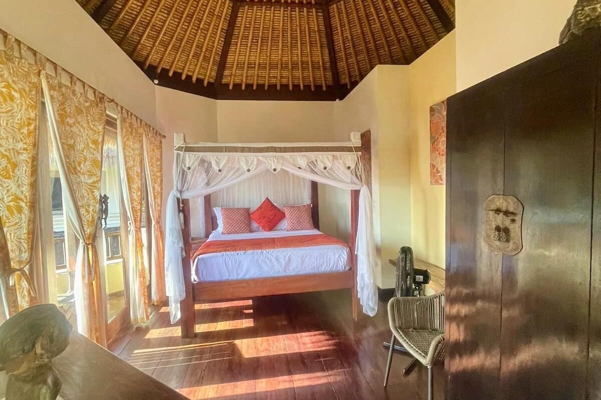 Quarto com cama sob telhado de palha, piso de madeira, porta preta e uma cadeira.