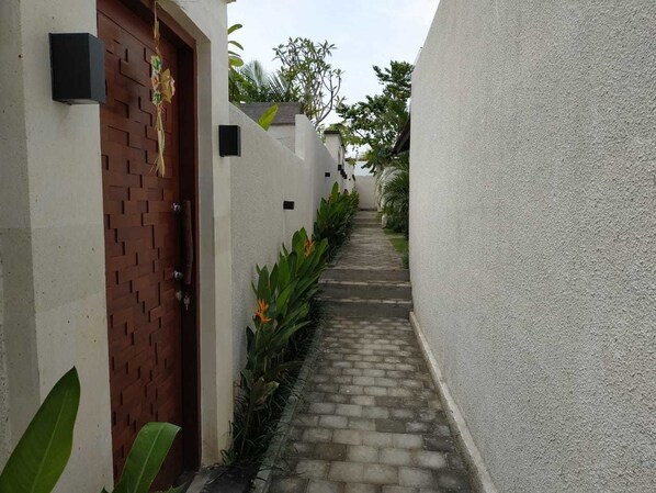 Un estrecho sendero que conduce a una puerta con diseño de ladrillo, flanqueado por paredes blancas y plantas verdes.