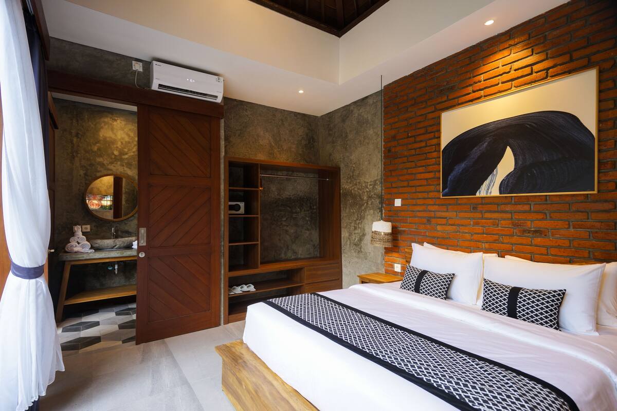 A modern bedroom with a large bed, a wooden wardrobe, a built-in TV, and a brick wall with a painting.