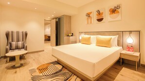 Una habitación de hotel moderna con una cama grande, un área de descanso con una silla y una mesa, y una mesita auxiliar con botellas.
