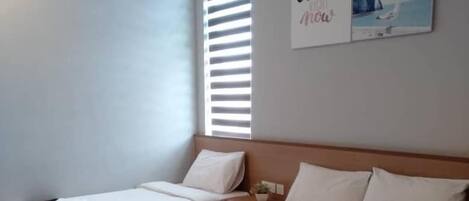 A hotel room with two beds, a large window with blinds, a wall-mounted artwork, and a small bedside table.