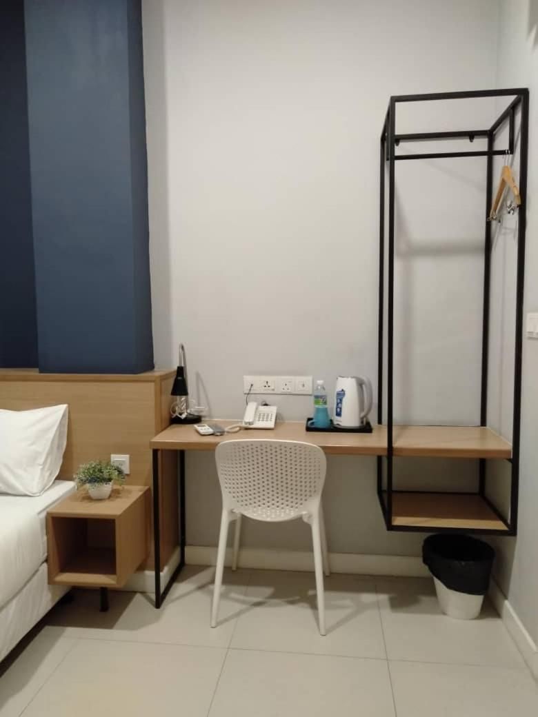 A small, compact hotel room with a bed, a desk, a chair, a telephone, a kettle, and a shelf.