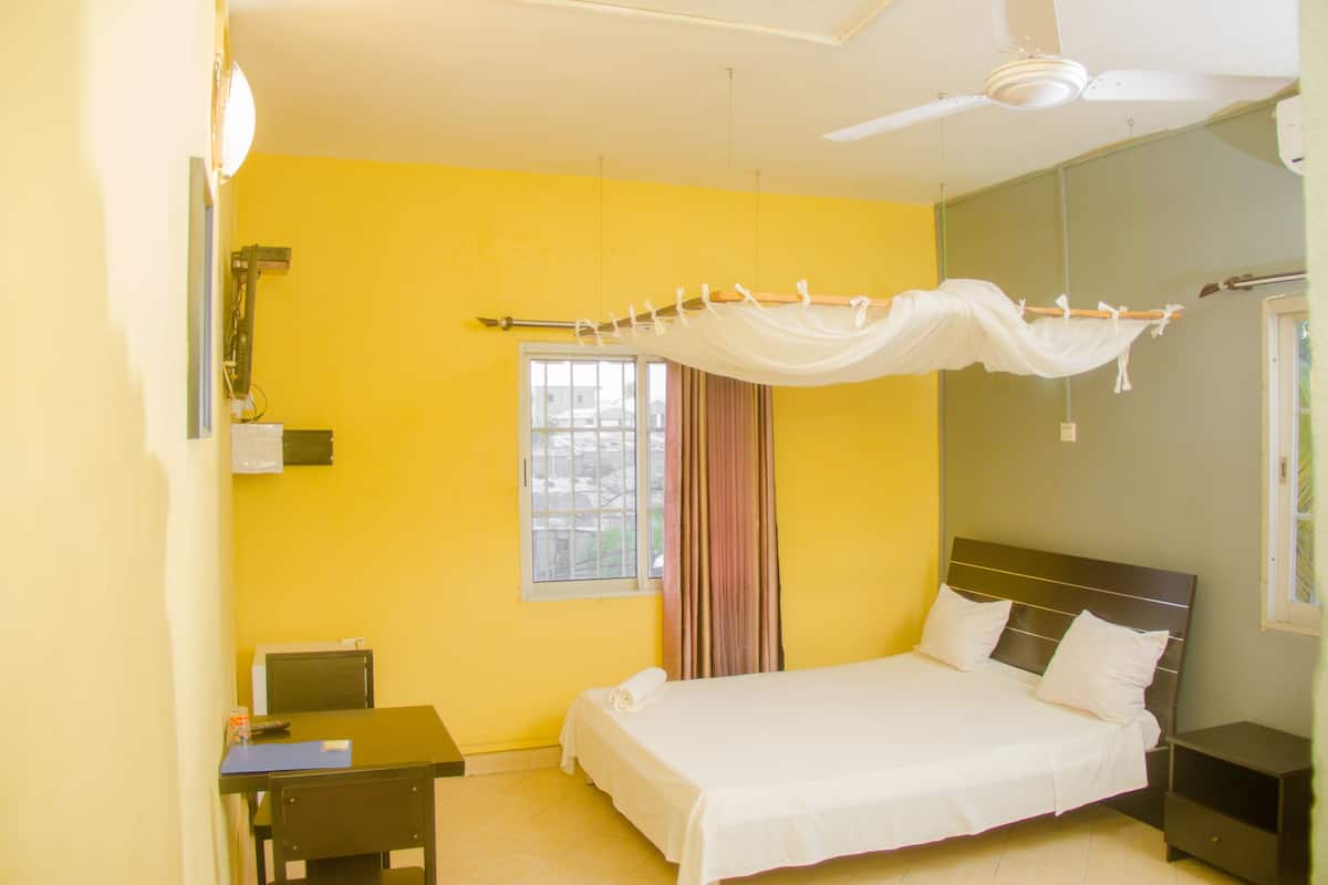 Ein Schlafzimmer mit Bett, Schreibtisch, Stuhl, Nachttisch, Deckenventilator, Fenster mit Vorhängen und einer Wandleuchte.