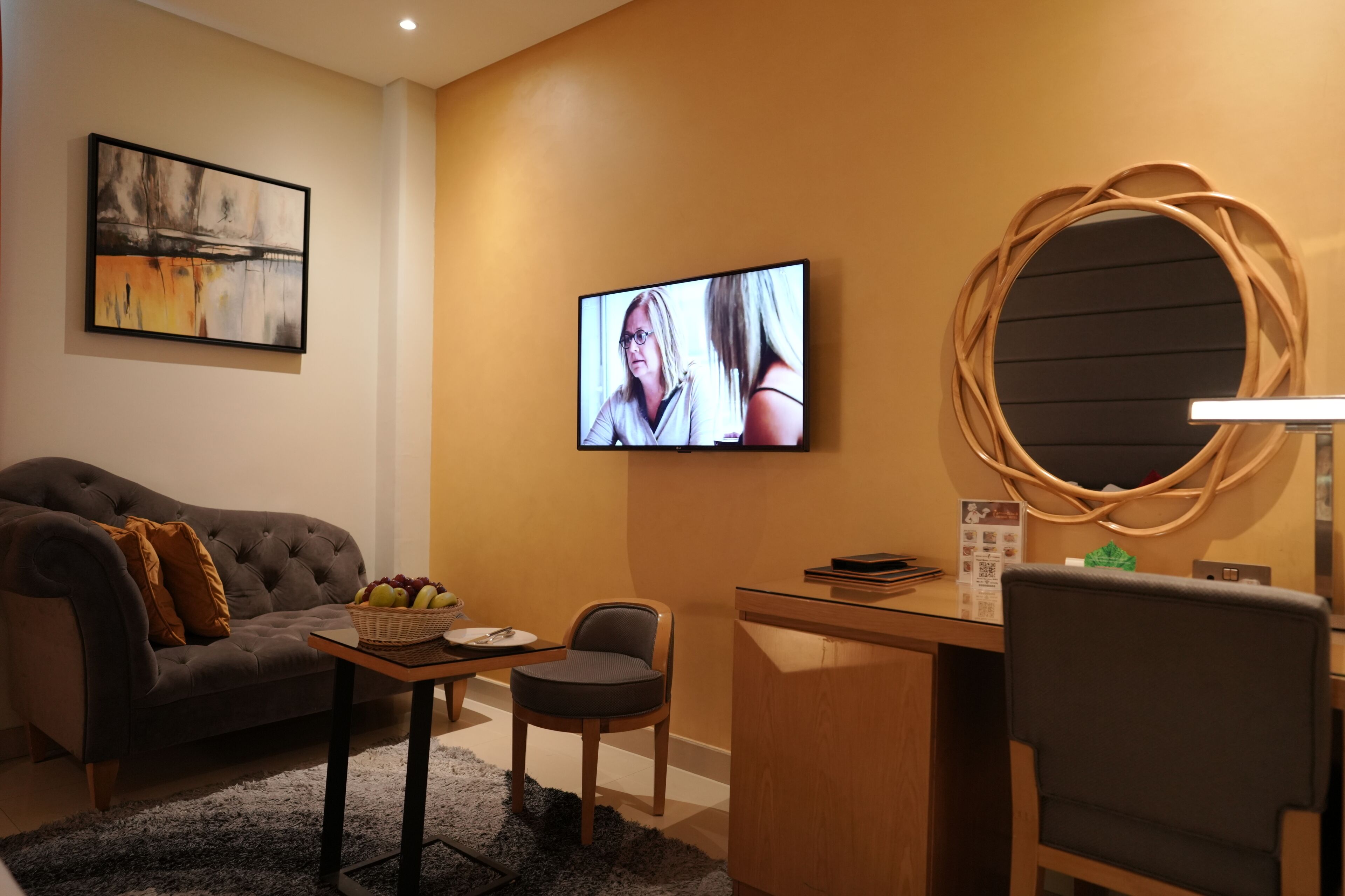 A modern hotel room with a grey sofa, a small wooden desk, a television, and a decorative mirror.