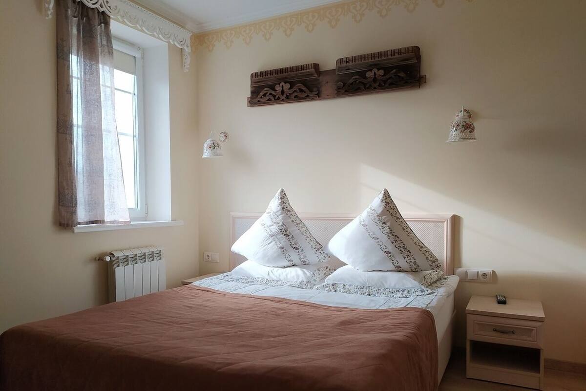 A neatly made bed with a brown bedspread, two decorative pillows, a bedside table with a lamp, and a window with curtains.
