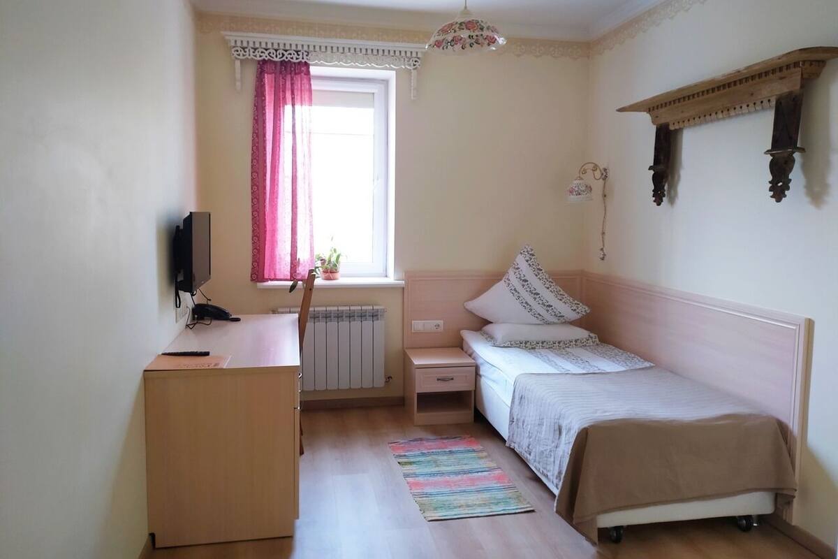 A bedroom with a bed, a desk, a chair, a radiator, a window with curtains, and a hanging light fixture.