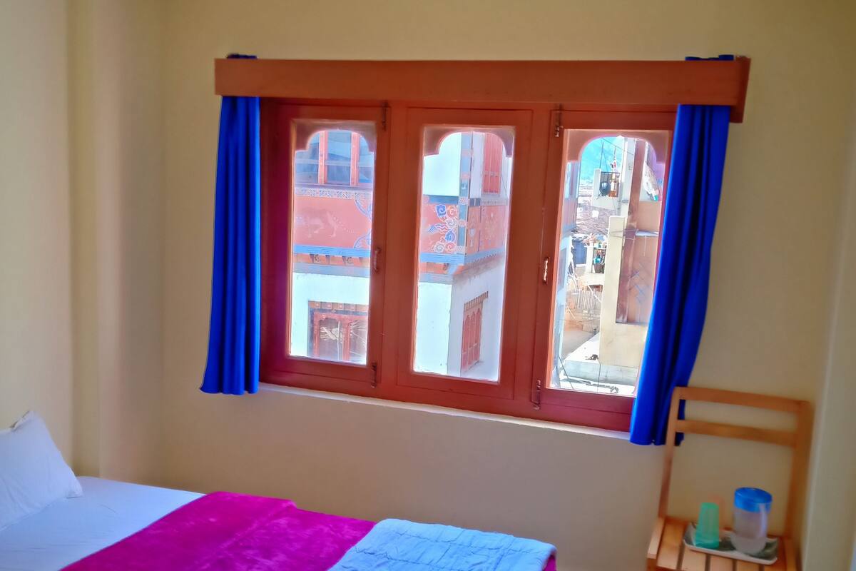 A room with a bed, a window with curtains, a small wooden table, and a view of buildings outside.