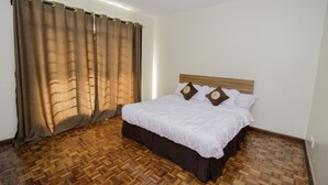 Quarto de hotel com piso de parquete de madeira, cama com roupa de cama branca e almofadas castanhas, e uma janela grande com cortinas marrons.