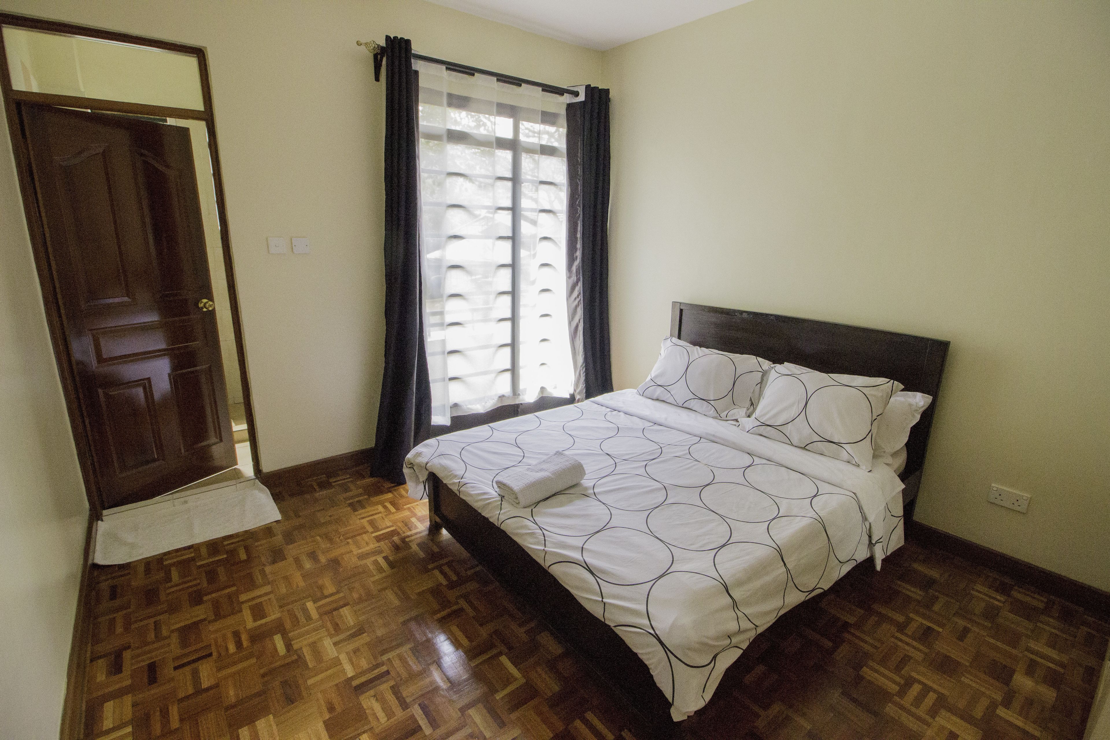 A bedroom with a wooden parquet floor, a bed with a white and black circular pattern bedspread, a dark headboard, and a window with black curtains.