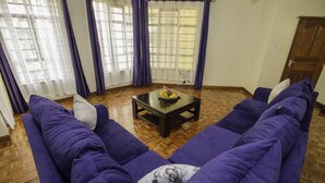 Uma sala de estar com dois sofás roxos, uma mesa de centro de madeira com uma tigela de frutas, e piso de madeira.