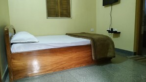 A single wooden bed with white bedding and a brown blanket, a television mounted on the wall, and a small shelf with a power outlet. - Nest Guest House (Bhubaneshwar)