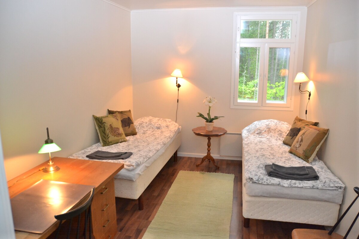 Une petite chambre accueillante, dotée de deux lits simples, d’un bureau en bois avec une lampe, d’une petite table avec une plante et d’une fenêtre donnant sur un paysage verdoyant.