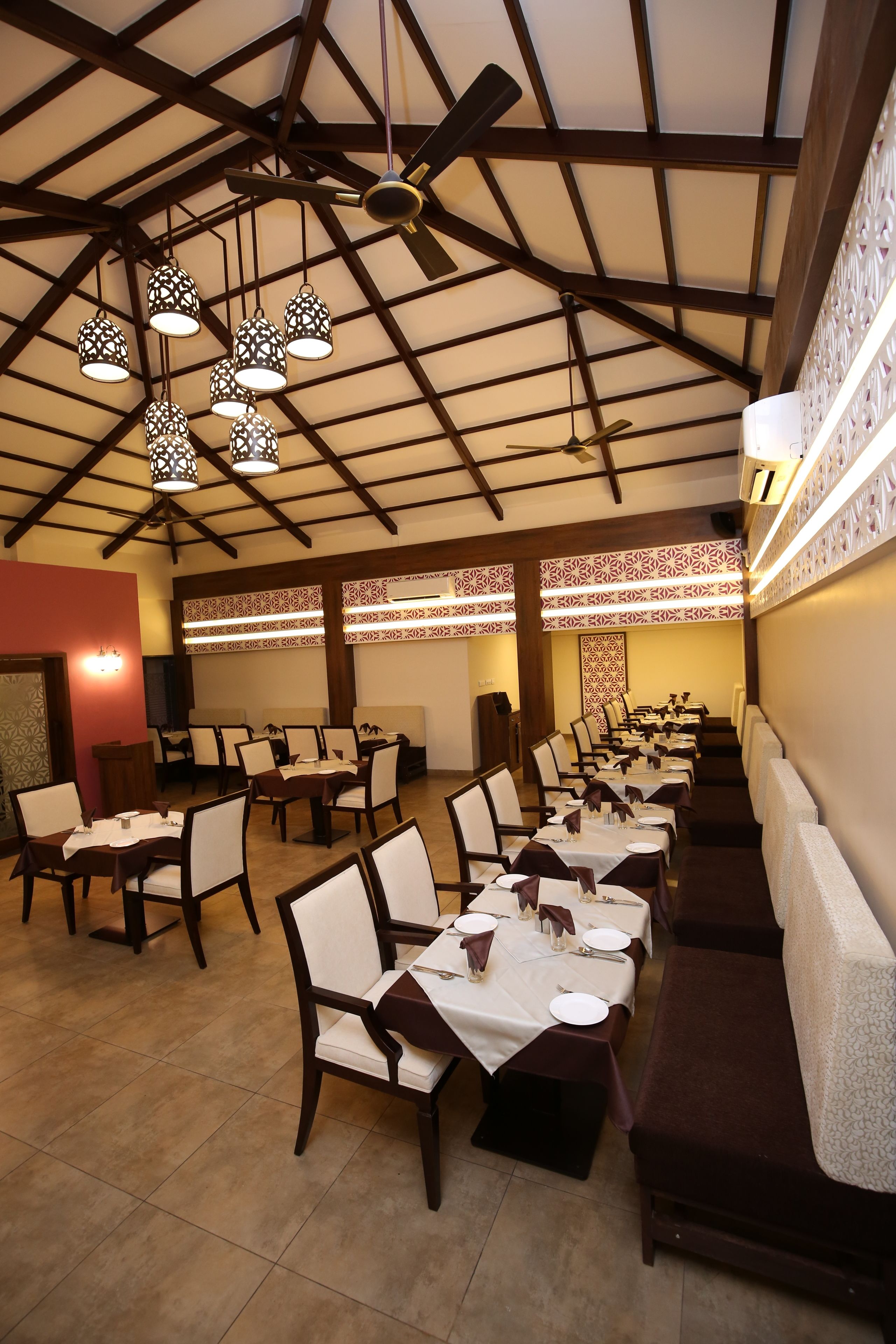 A restaurant with a high ceiling, exposed beams, and patterned wall designs. Tables are set with white tablecloths and dark chairs.