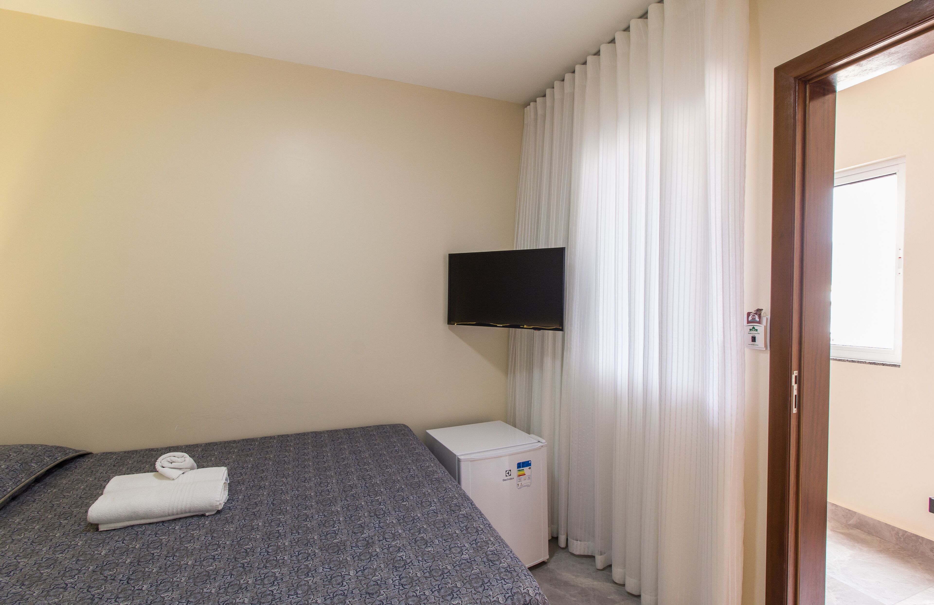 A hotel room with a bed, a small white refrigerator, a flat-screen TV mounted on the wall, and a door with a window.