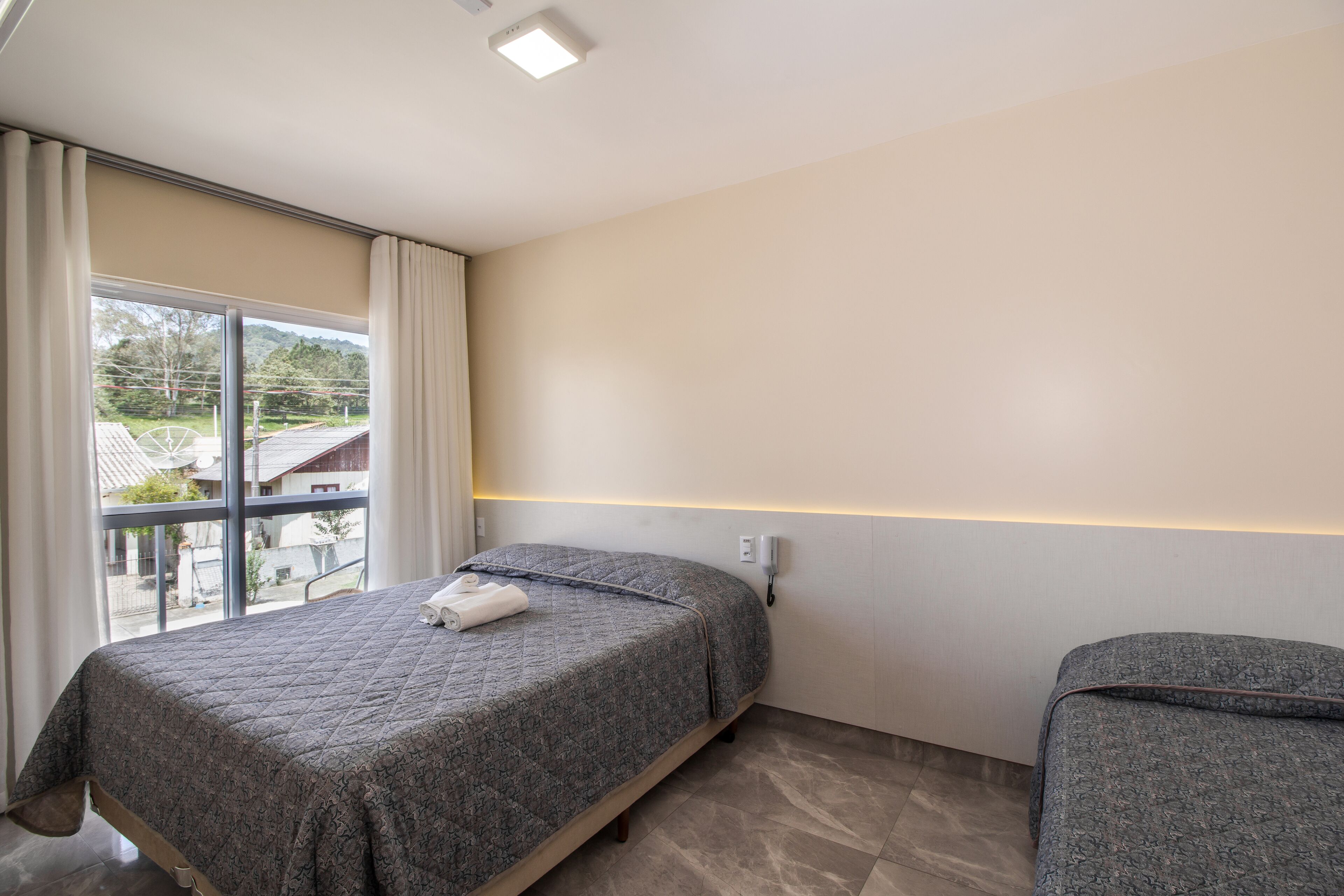 A bedroom with a bed, a bedside table, a window with curtains, and a view of the outside.