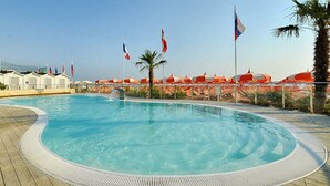 A swimming pool with a curved edge, surrounded by a deck and palm trees. Flags are visible in the background. - Hotel Quisisana Riccione (Riccione)