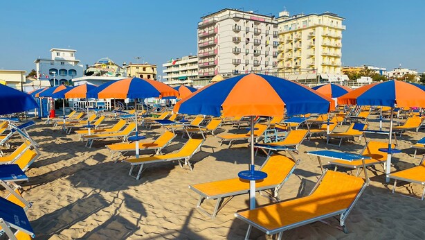 A beach with rows of sun loungers and umbrellas, a clear sky, and buildings in the background. - Hotel Quisisana Riccione (Riccione)