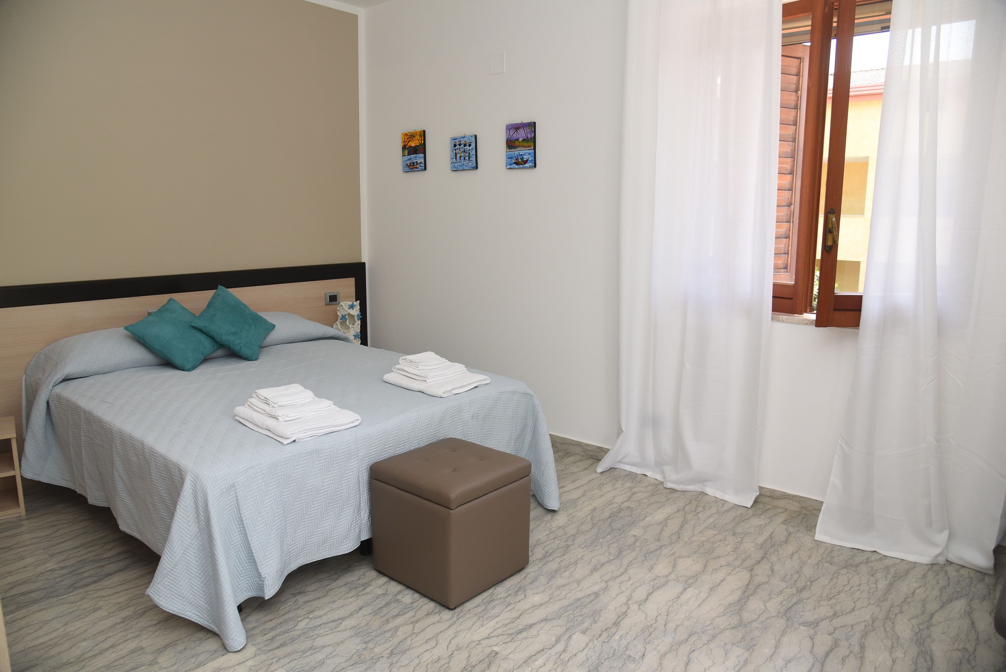 A neatly made bed with a light blue bedspread and teal pillows, a small brown ottoman, and three framed artworks on the wall.