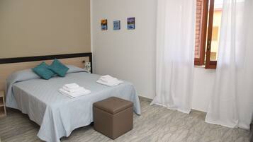A neatly made bed with a light blue bedspread and teal pillows, a small brown ottoman, and three framed artworks on the wall.