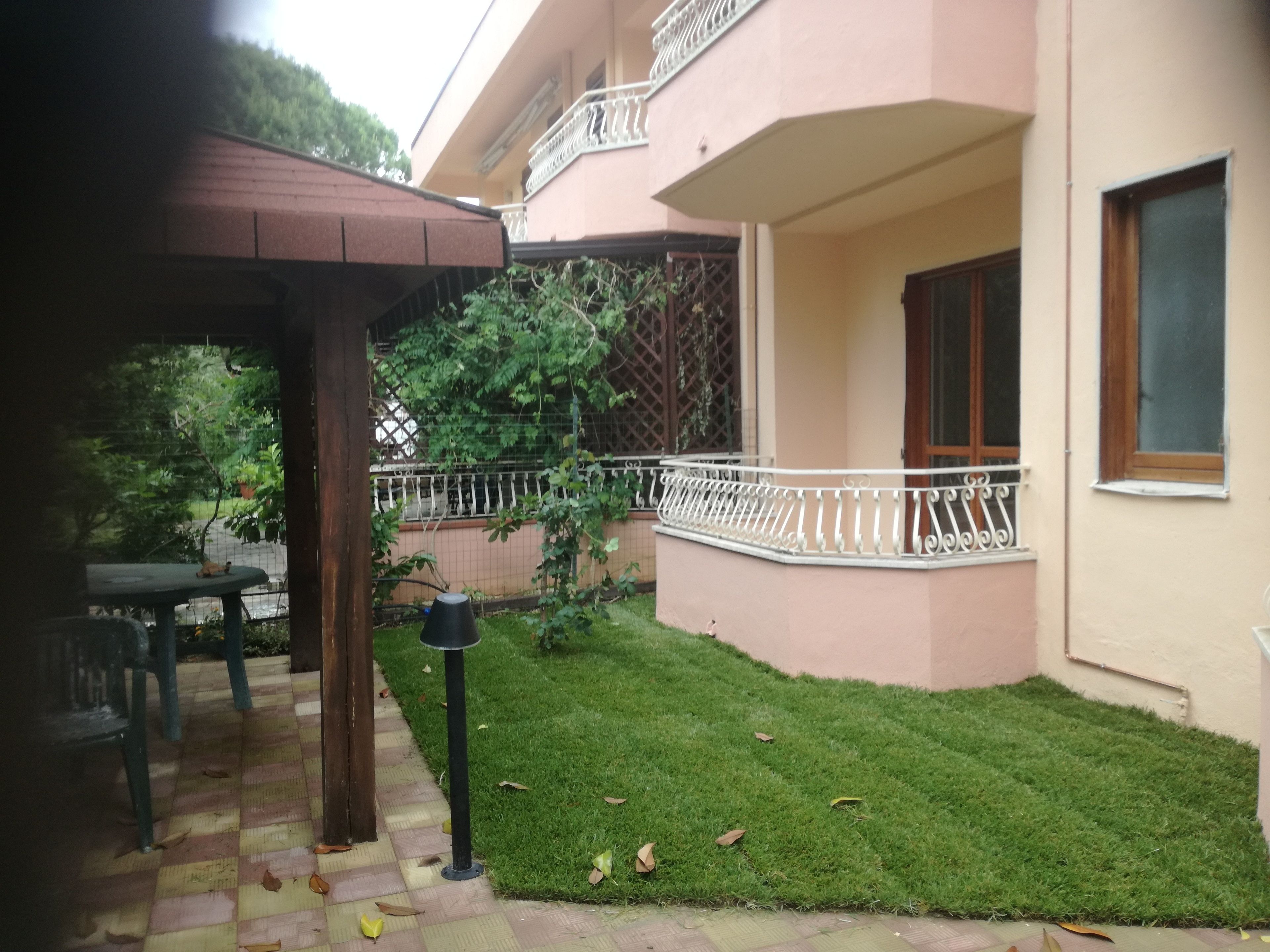Um edifício residencial com um gramado bem cuidado, uma pérgula de madeira e uma varanda com guarda-corpo branco.