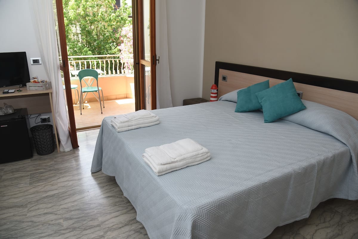 Habitación de hotel con cama, escritorio con televisor y balcón con silla.