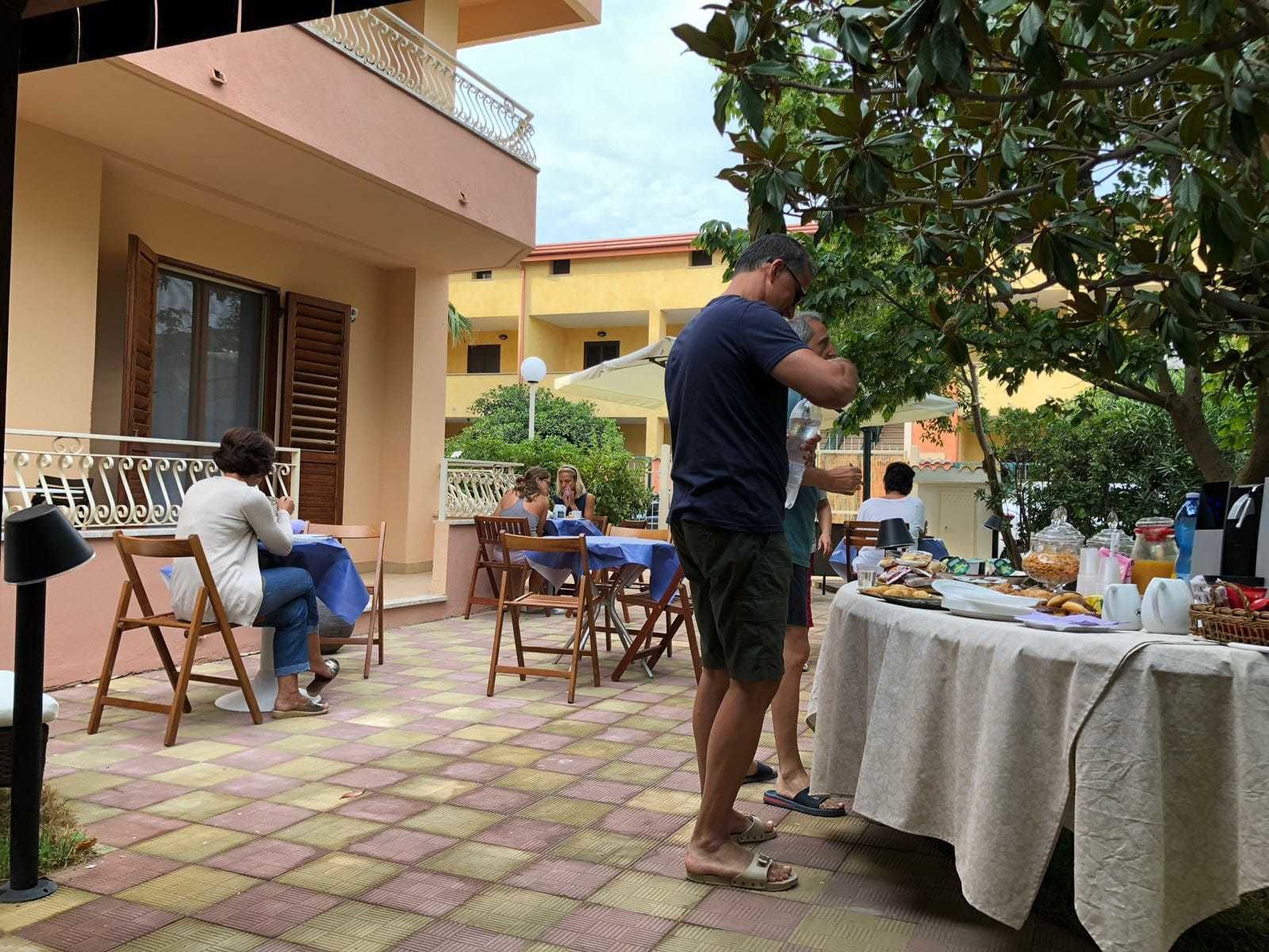 Un área de comedor al aire libre con mesas y sillas, una mesa de buffet con comida y bebidas, y un edificio con balcones al fondo.