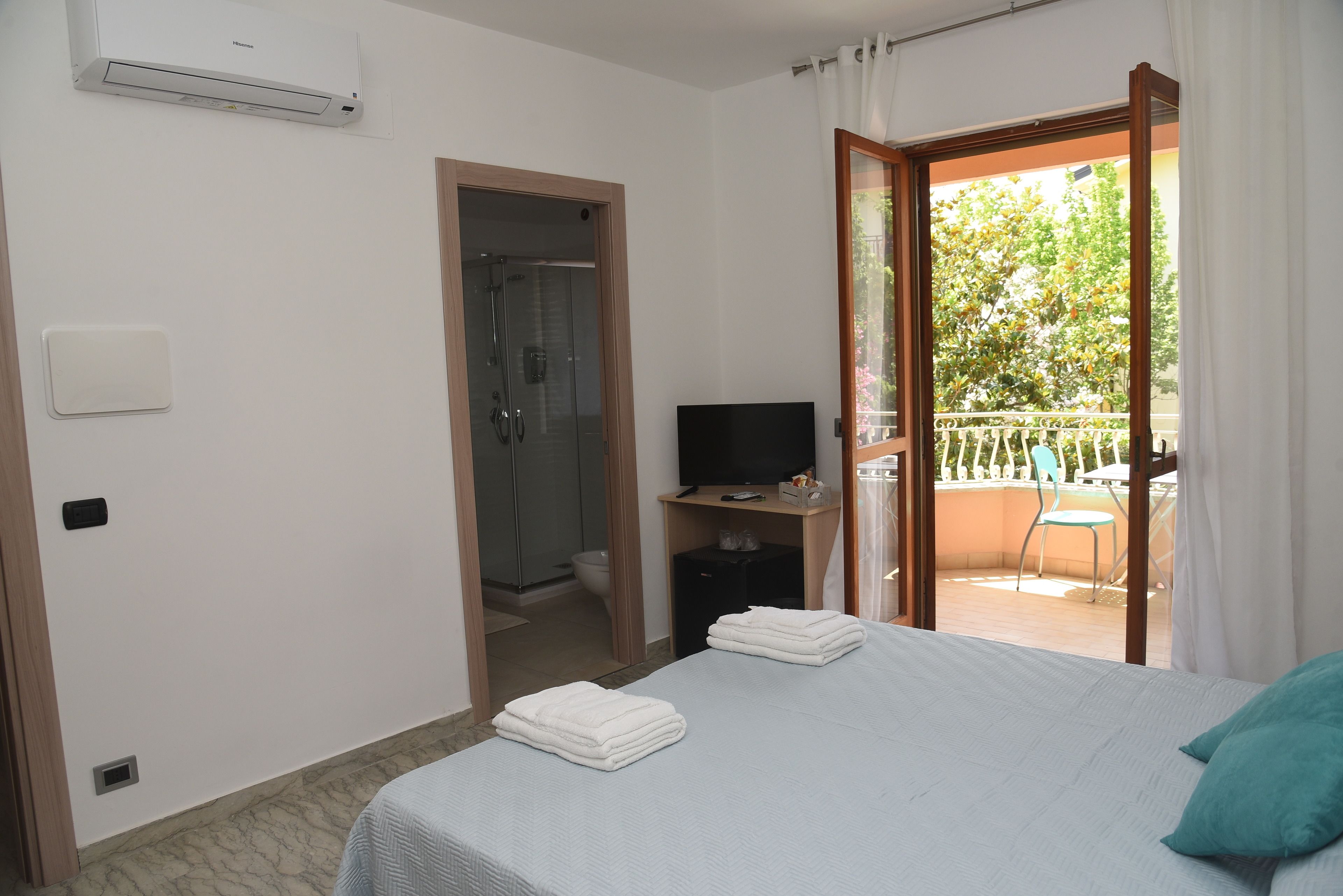 A hotel room with a bed, a TV, a shower, and a balcony with a chair and table.
