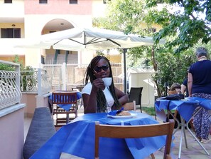 Un'area pranzo all'aperto con tavoli ricoperti da tovaglie blu, ombrelloni bianchi e un edificio con balconi sullo sfondo.