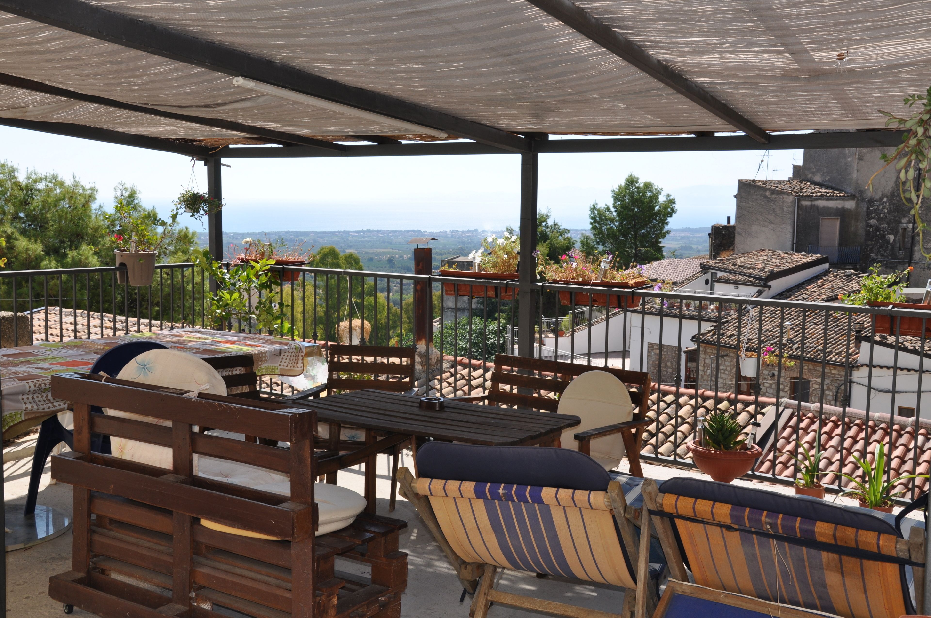 A covered outdoor seating area with wooden furniture and potted plants, overlooking a cityscape.