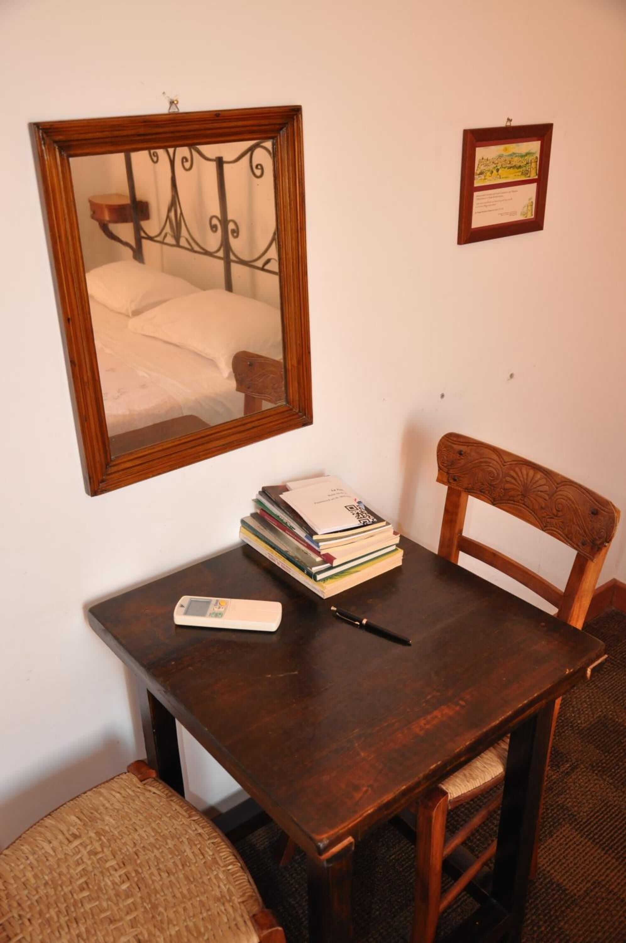 Ein kleiner Raum mit einem Holztisch, einem Stuhl, einem Bücherstapel, einer Fernbedienung und einem gerahmten Bild an der Wand.