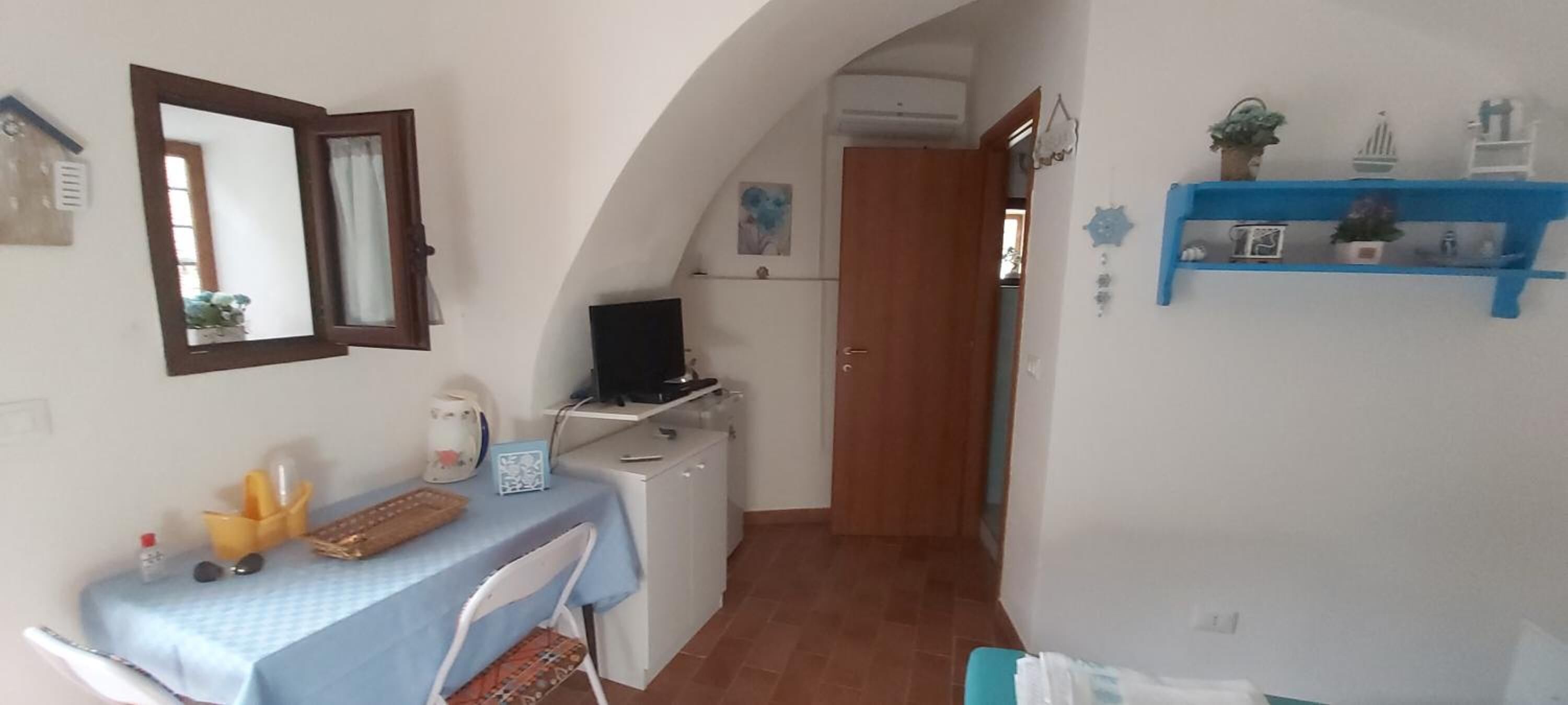 A compact kitchen with a dining area, a TV on a stand, and a blue shelf with decorative items.