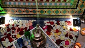 A tiled ceiling with geometric patterns and a water spout. Below, a table is covered with various fruits and a teapot. - Riad Dar Leuleu (Marrakech)