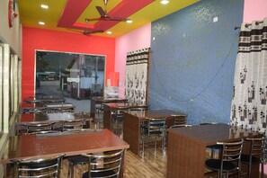 A restaurant with multiple tables and chairs, a ceiling fan, and a window with a view of the street. - Bagori Resort (Golaghat)