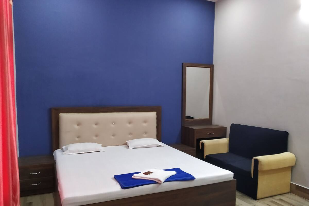 Una habitación de hotel con cama, sillón, mesita de noche, espejo y suelo de madera.