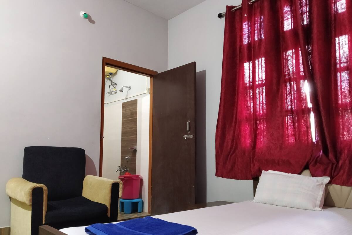 Habitación de hotel con una cama, una silla, un taburete pequeño, una cortina roja y una puerta que da a un baño.
