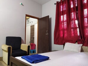A hotel room with a bed, a chair, a small stool, a red curtain, and a door leading to a bathroom. - Bagori Resort (Golaghat)