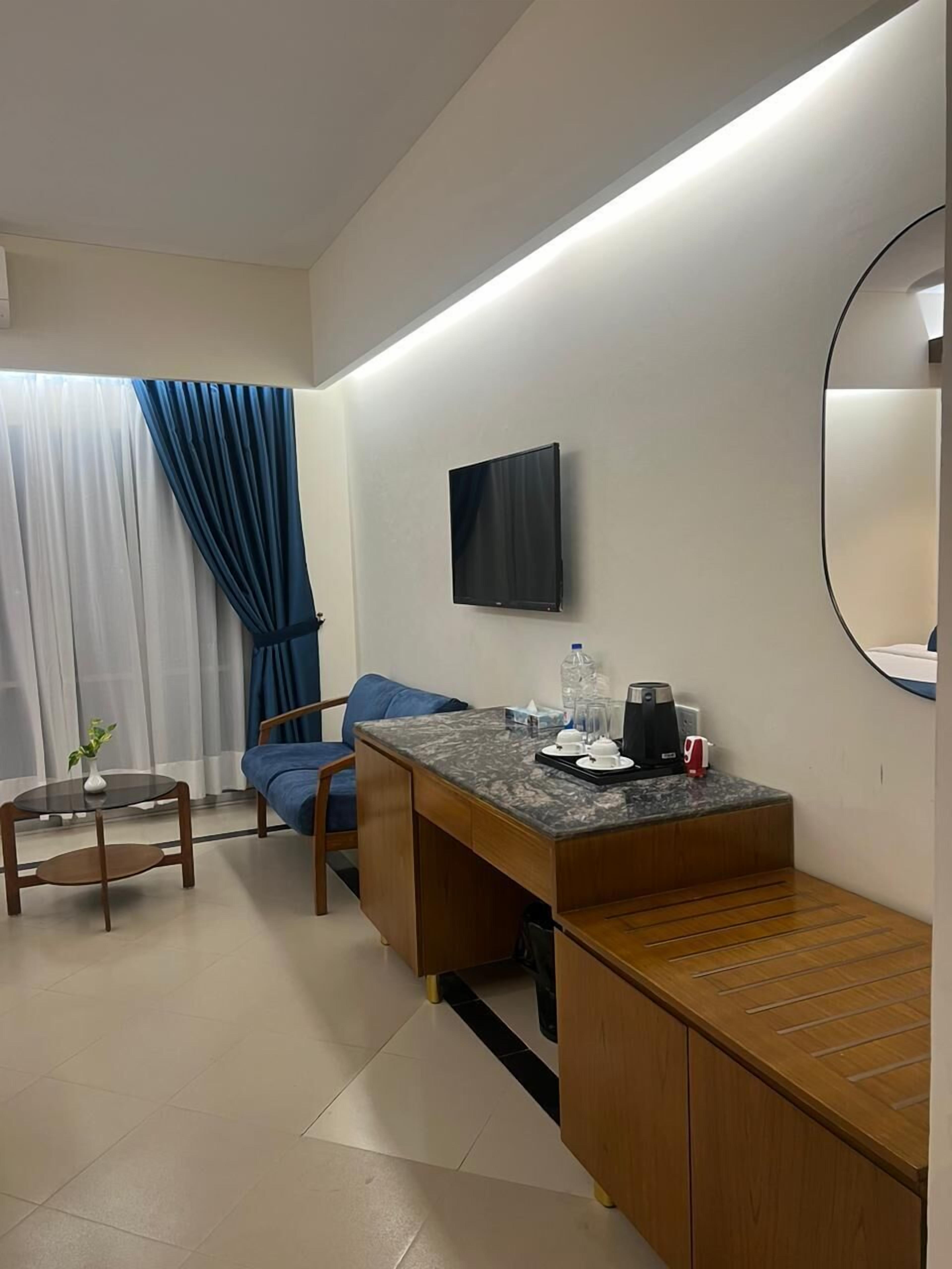 Ein modernes Hotelzimmer mit Schreibtisch aus Holz, einem blauen Sofa, einem kleinen runden Tisch mit einer Vase, einem flachen Fernseher an der Wand und einem großen Spiegel.
