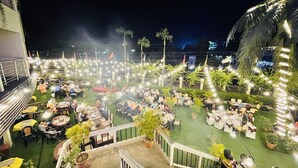 Un espacio de comedor al aire libre con mesas y sillas, rodeado de follaje e iluminación con luces colgantes.