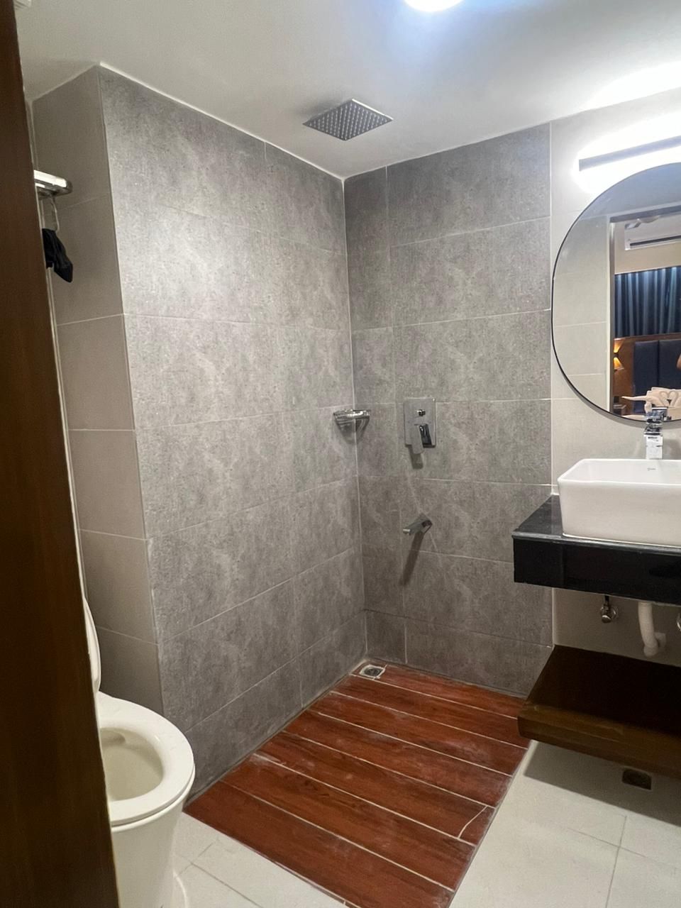 A bathroom with grey tiles, a wooden floor, a white toilet, a sink with a mirror, and a shower.