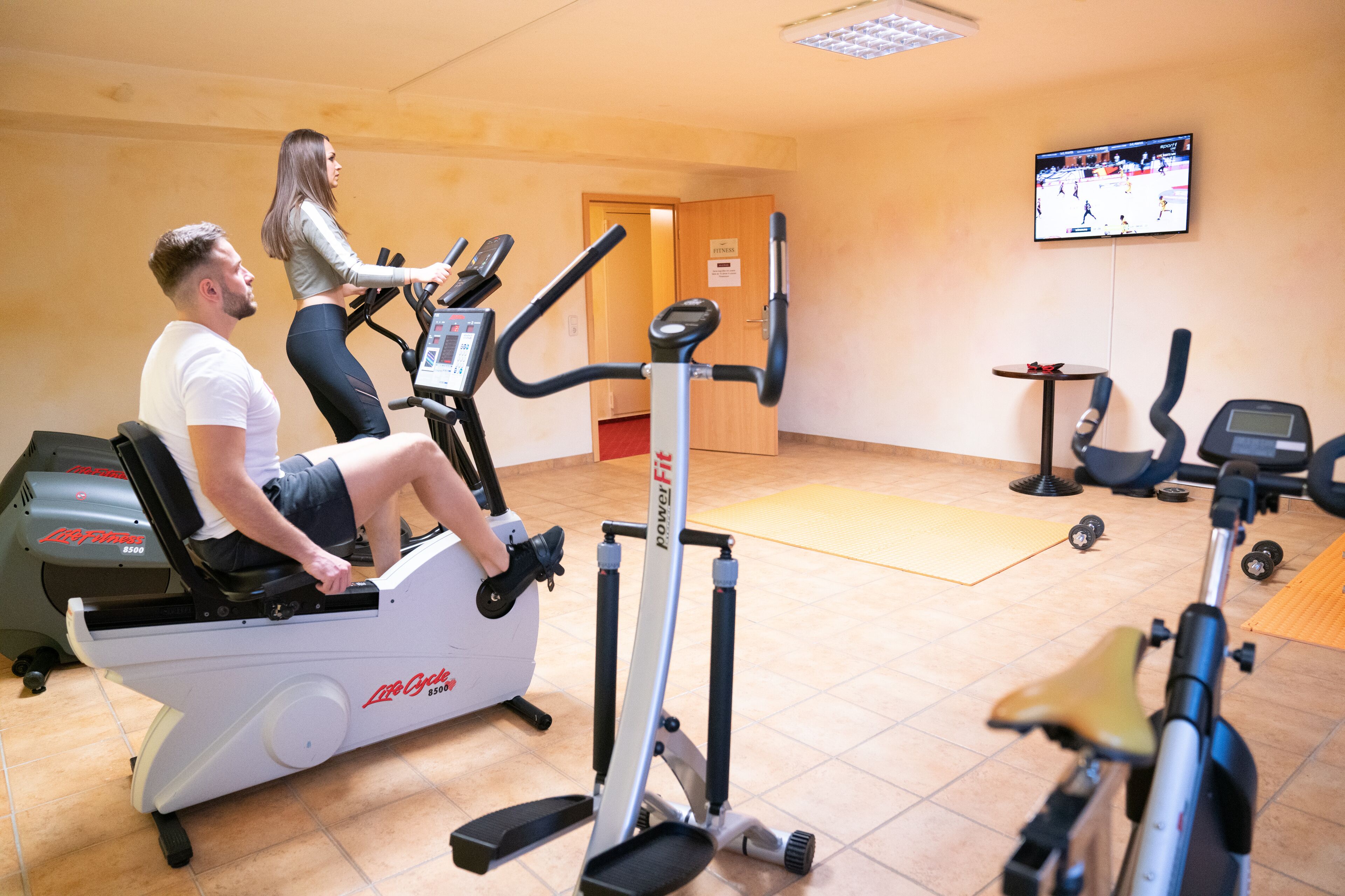 Une salle de sport équipée de vélos d’appartement, d’un tapis de course et d’un téléviseur diffusant un événement sportif.