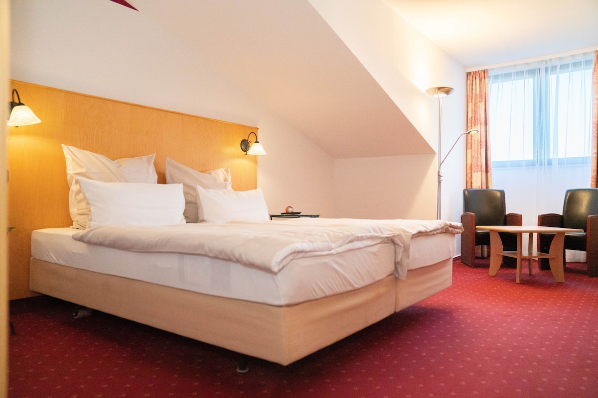 Habitación de hotel con una cama grande, dos lámparas de noche, una mesita, dos sillas y un ventanal con cortinas.