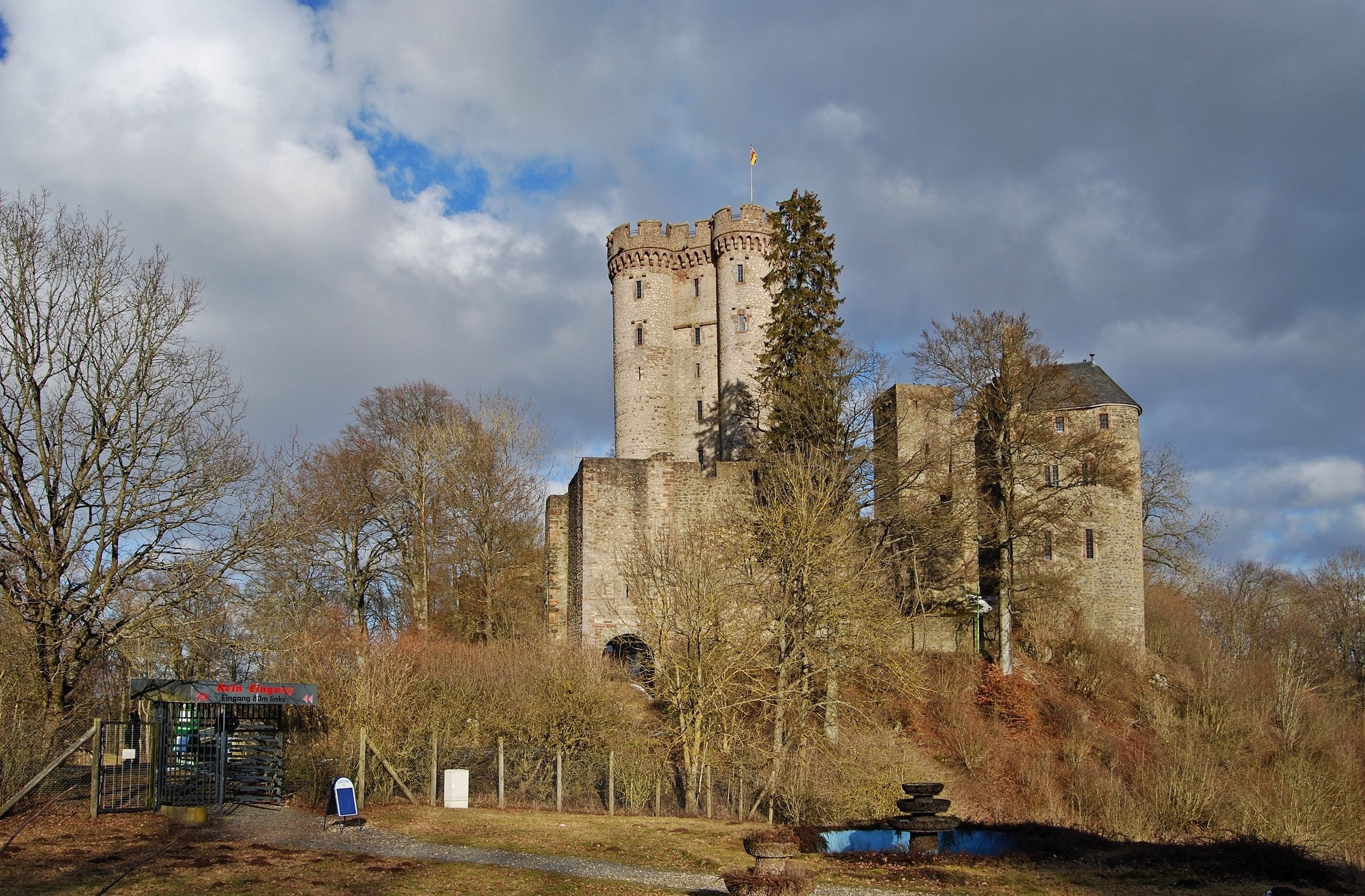 A historic stone castle with a tall tower, surrounded by trees and a small gatehouse.