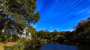 A riverside view with a white building featuring a balcony, surrounded by lush greenery and trees. - Short Walk to Beach. Dog-Friendly, Renovated, Bright & Modern Villa with Lagoon Views. (Kiawah Island)