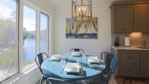 A dining area with a glass table, chairs, and a view of a lake through large windows. - Short Walk to Beach. Dog-Friendly, Renovated, Bright & Modern Villa with Lagoon Views. (Kiawah Island)